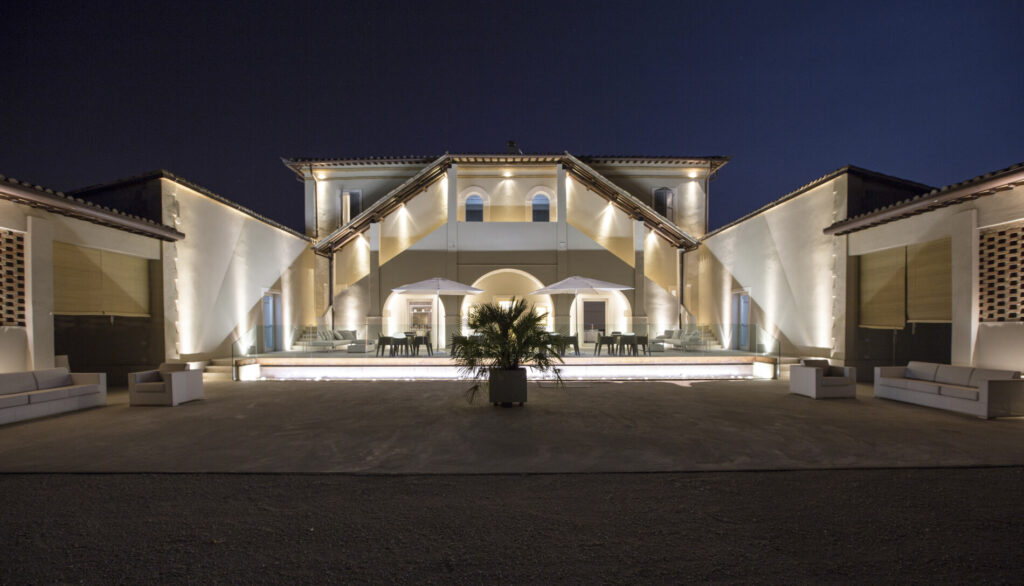 Cortile Le Colombare di notte Cortile a U dell'Hotel Le Colombare di notte, con facciate illuminate, terrazza con tavoli e ombrelloni bianchi e divani lounge in primo piano