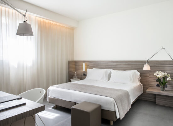Camera Classic moderna con letto matrimoniale, pannello in legno a parete, scrivania bianca e pouf grigio ai piedi del letto