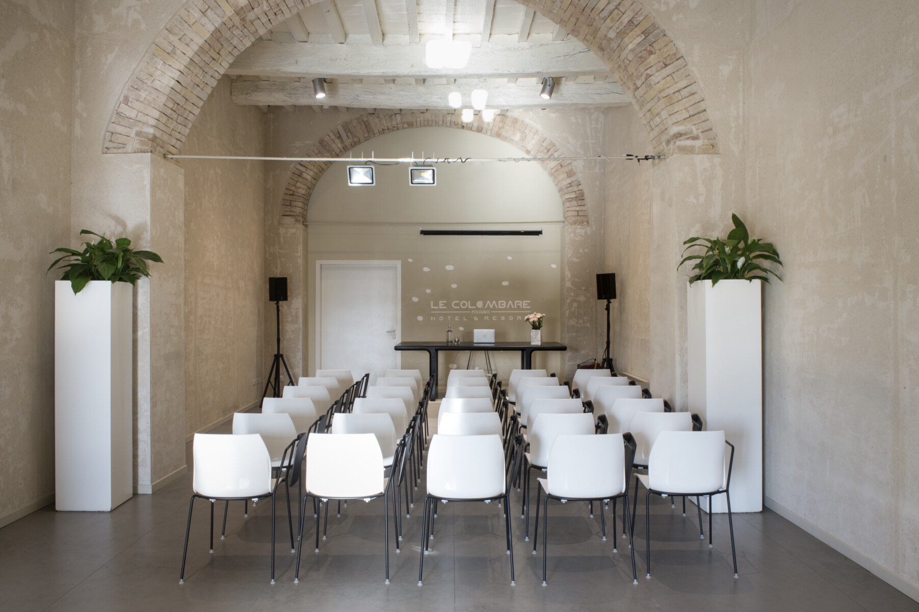 Sala convegni con file di sedie bianche, travi in legno e archi in mattoni; sul fondo il logo Hotel Le Colombare Foligno