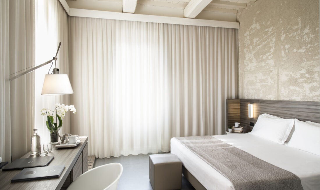 Camera Superior con travi a vista, letto con copriletto grigio, pouf ai piedi e scrivania con orchidee e tende velate