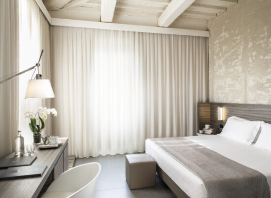 Camera Superior con travi a vista, letto con copriletto grigio, pouf ai piedi e scrivania con orchidee e tende velate