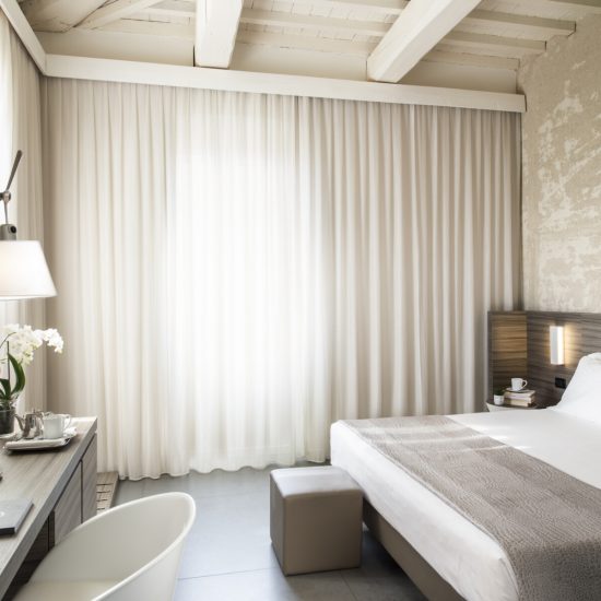 Camera Superior con travi a vista, letto con copriletto grigio, pouf ai piedi e scrivania con orchidee e tende velate