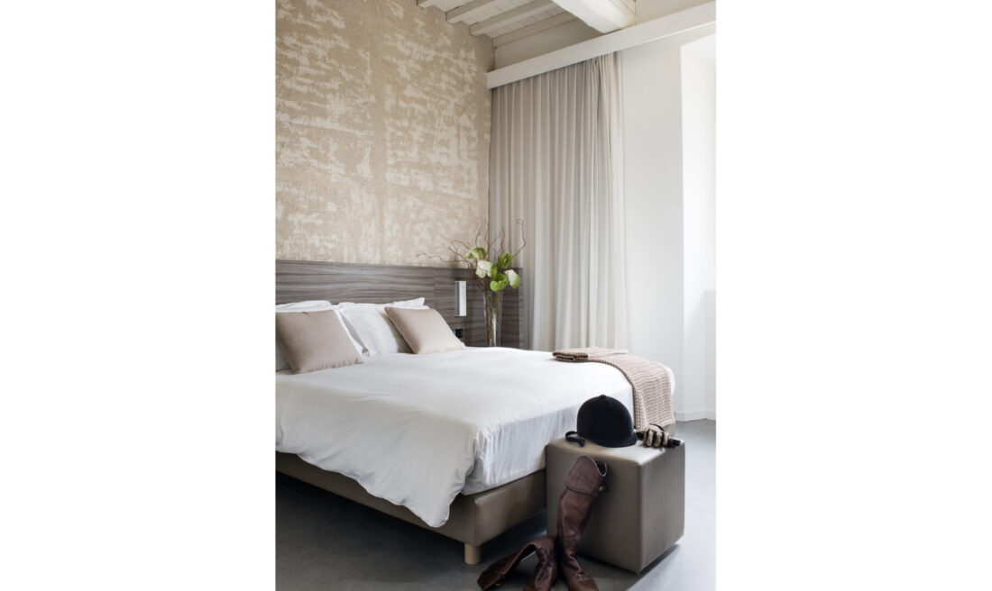 Dettaglio camera Superior con parete materica, travi bianche, letto bianco e stivali da equitazione su pouf in pelle