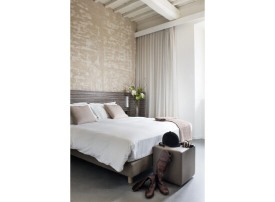 Dettaglio camera Superior con parete materica, travi bianche, letto bianco e stivali da equitazione su pouf in pelle