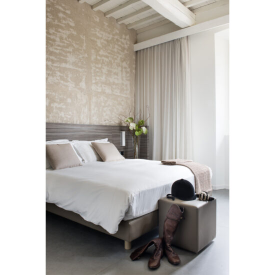 Dettaglio camera Superior con parete materica, travi bianche, letto bianco e stivali da equitazione su pouf in pelle