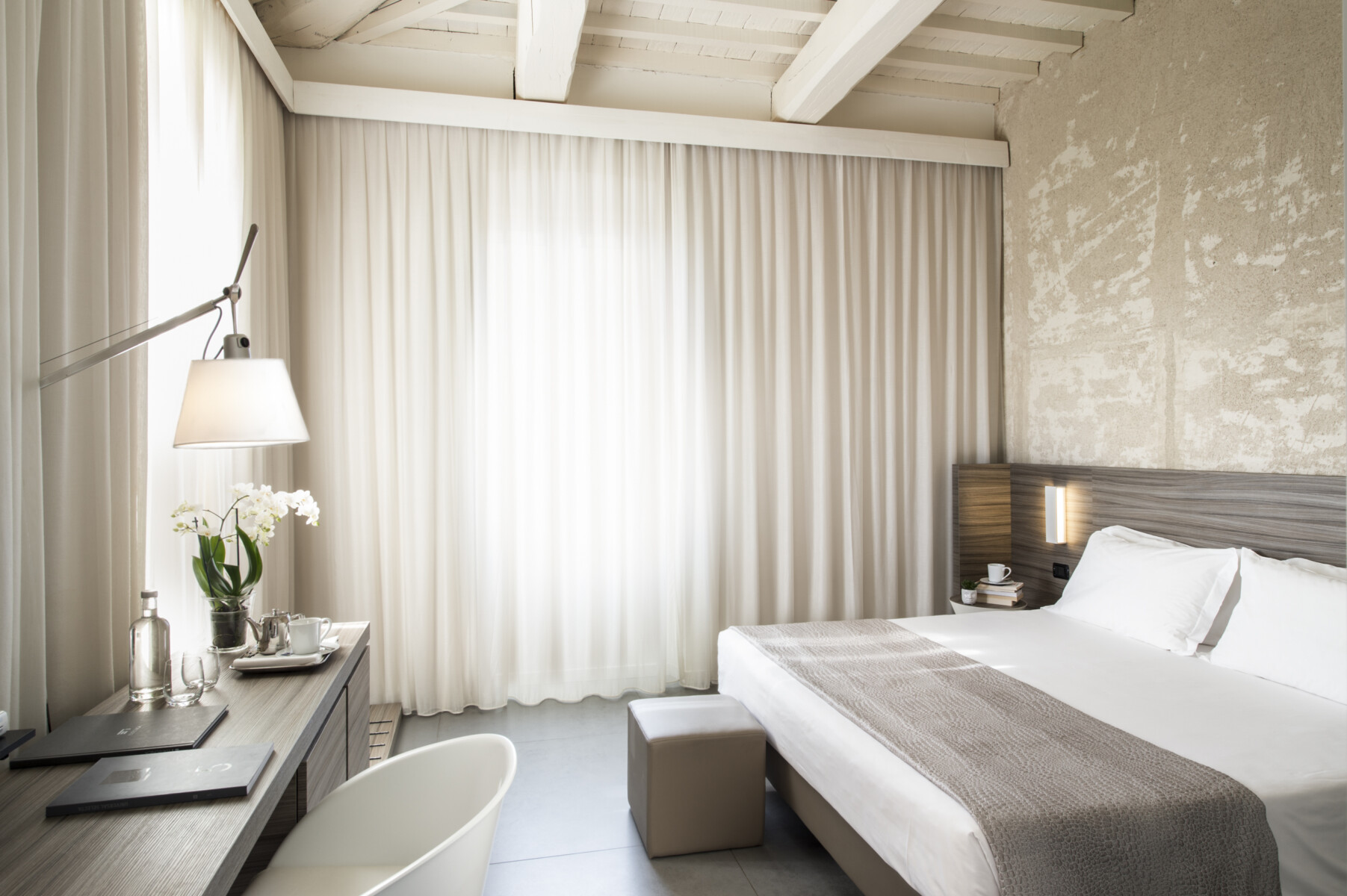 Camera Superior con travi a vista, letto con copriletto grigio, pouf ai piedi e scrivania con orchidee e tende velate