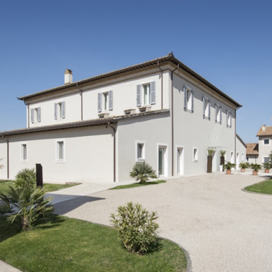 Facciata esterna a due piani dell'Hotel Le Colombare con tetto in coppi, persiane grigie e vialetto ghiaioso fiancheggiato da prato e palme