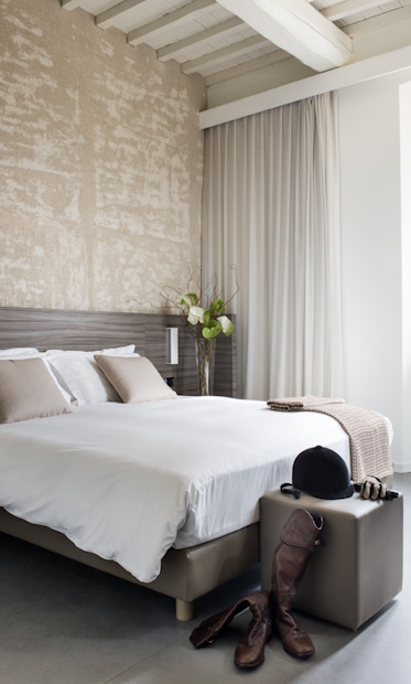 Camera Superior con letto matrimoniale, testata in pietra, soffitto con travi bianche, fiori di calla e equipaggiamento da equitazione sul pouf