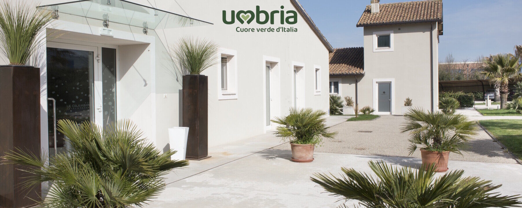 Hero ingresso Le Colombare Ingresso dell'Hotel Le Colombare con facciata bianca, logo Umbria Cuore verde d'Italia, vialetto in pietra e vasi con palme