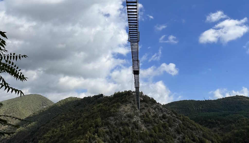 Ponte tibetano di Sellano visto dal basso: passerella metallica sospesa sulla valle verde verso le colline sotto un cielo azzurro con nuvole