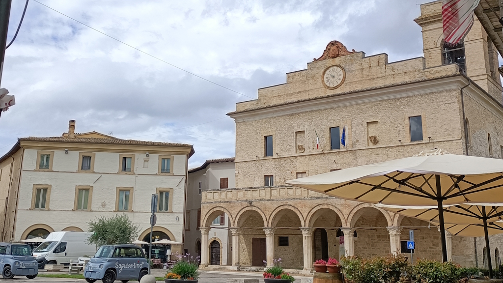 Piazza di Montefalco con palazzo comunale a loggia, torre campanaria e due microcar elettriche con scritta Sagrantino sotto un cielo nuvoloso