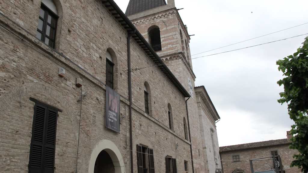 Edificio in pietra di Spello con banner "Pinacoteca Civica e Diocesana Spello" appeso alla finestra e campanile con orologio sullo sfondo